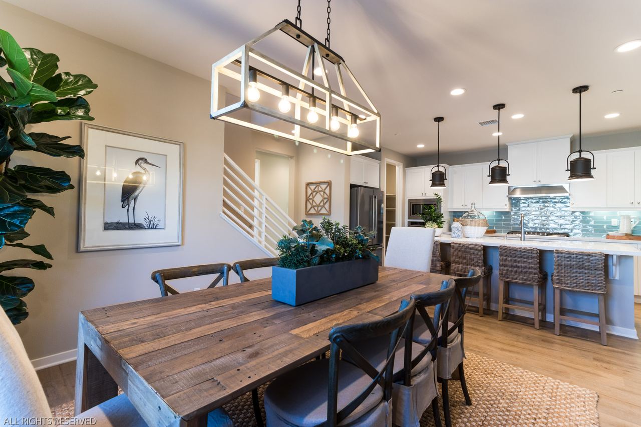 Farmhouse dining table under a linear chandelier, opening to a bright kitchen